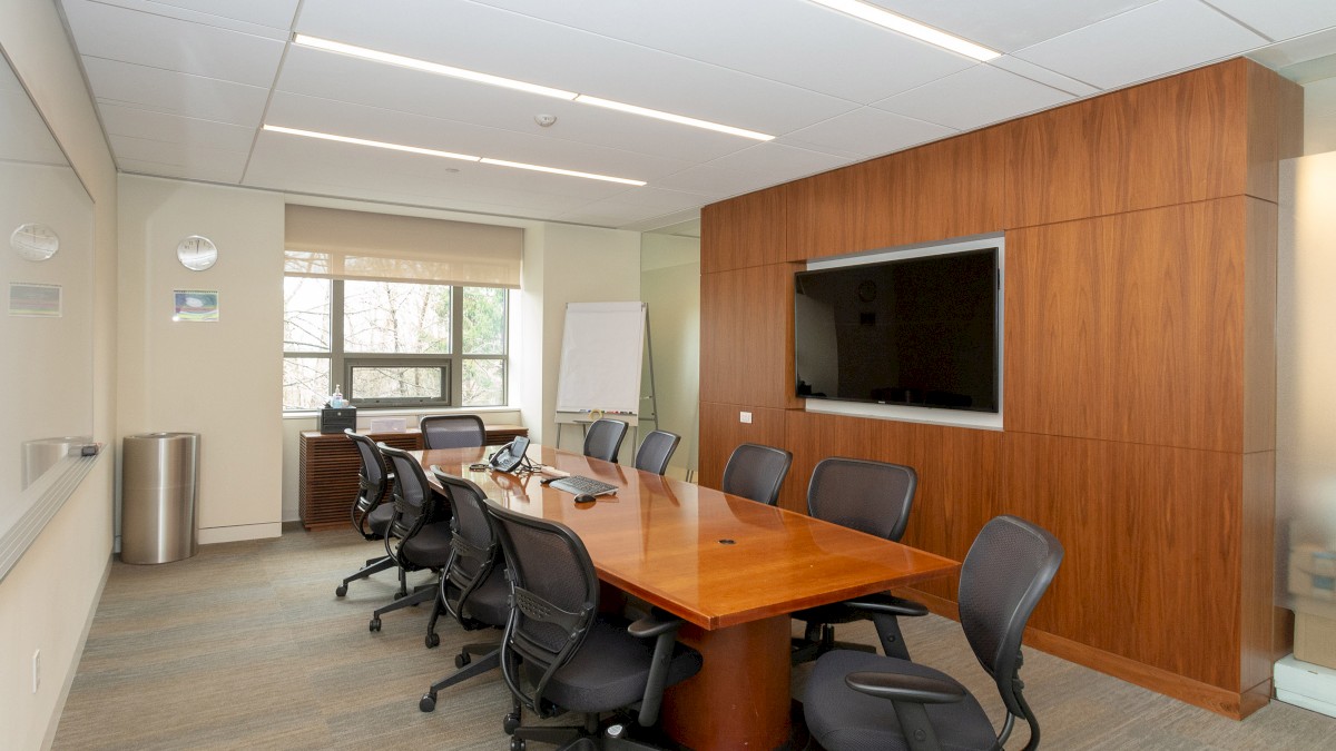 A modern conference room with a large wooden table, multiple office chairs, a whiteboard, a wall-mounted TV, and a window.