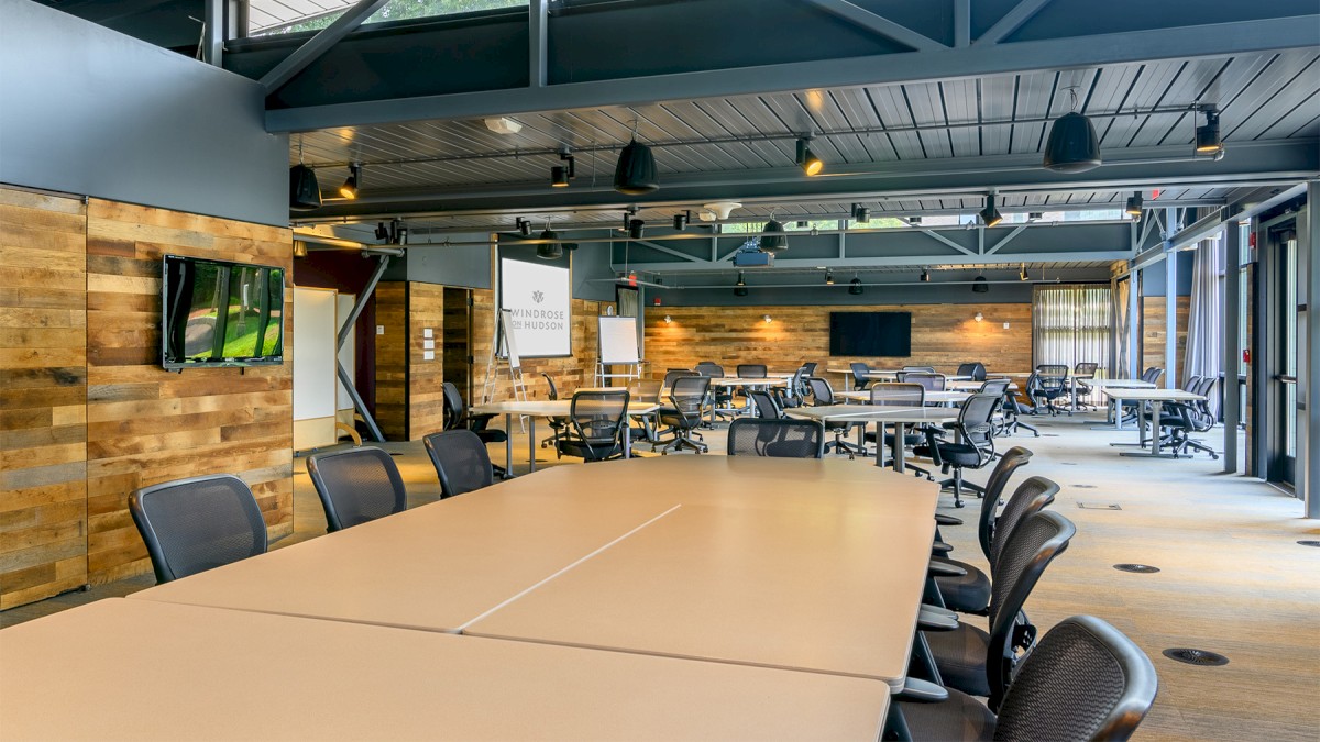 A modern conference room with wood accents, large tables, multiple chairs, and screens, illuminated by skylights and track lighting.