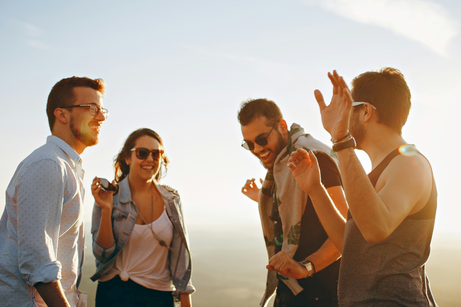 A group of four people, three men and one woman, are outdoors laughing and having a good time, with a clear sky in the background.