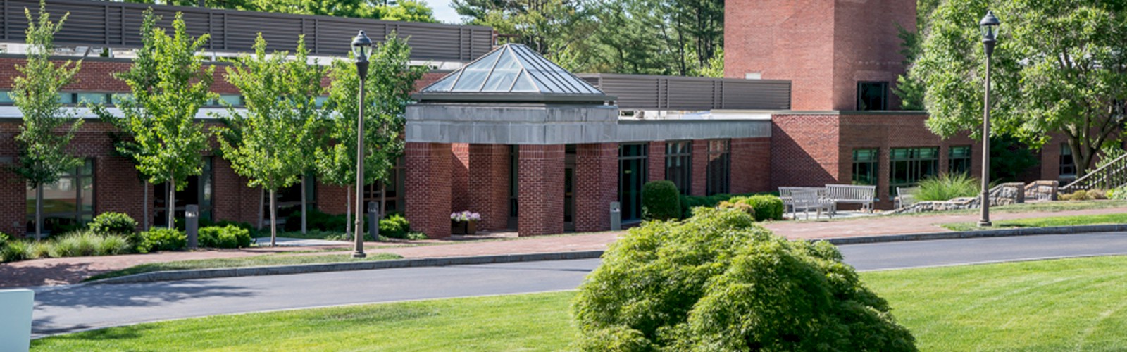 A brick building surrounded by green grass, trees, and shrubs, under a partly cloudy sky, with a driveway running in front and street lamps along the path.
