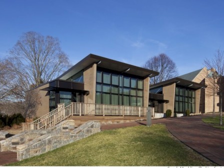 The image shows a modern building with large glass windows, a stone-paved walkway, and a sloped roof, surrounded by trees and grass.