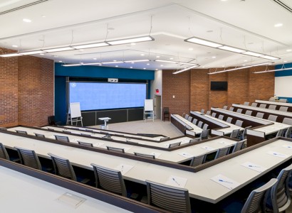 A modern lecture hall with tiered seating, a large screen at the front, whiteboards, and desks equipped with notepads and pens, set for a presentation.