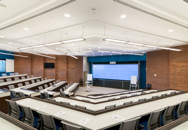 This image shows an empty lecture hall with tiered seating, tables, a large screen, and brick walls, ready for a presentation or class.