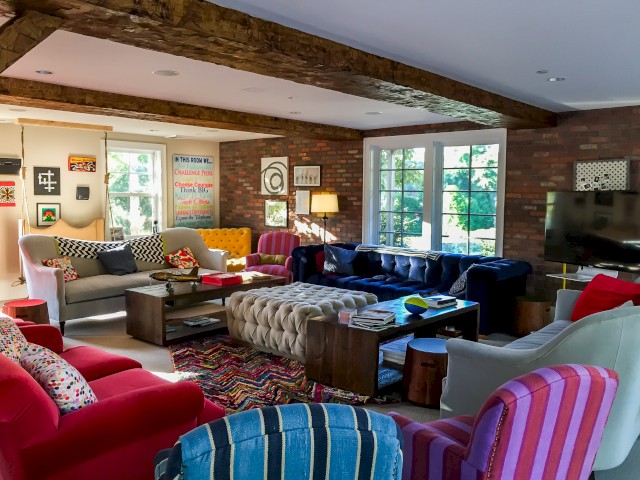 A vibrant living room features colorful furniture, a tufted ottoman, exposed brick walls, wooden beams, large windows, and eclectic artwork.