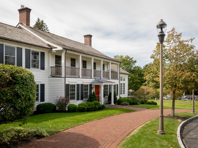 A white two-story house with black shutters, a red door, a brick walkway, and landscaped greenery. It is a bright, sunny day with a lamppost nearby.