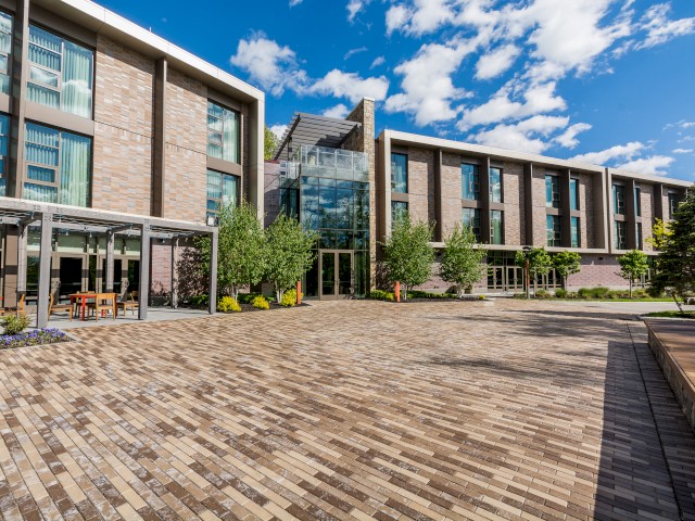 A modern building with large windows and an extensive brick courtyard, surrounded by trees and outdoor seating, is shown.