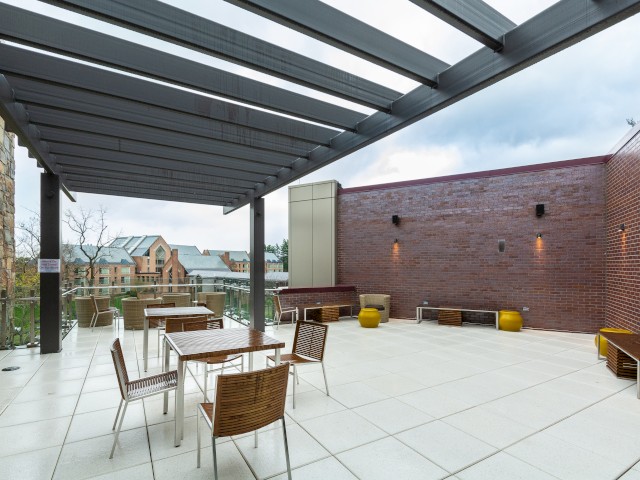 An outdoor patio with a pergola, wooden tables, chairs, yellow stools, and brick walls, overlooking buildings in the background.