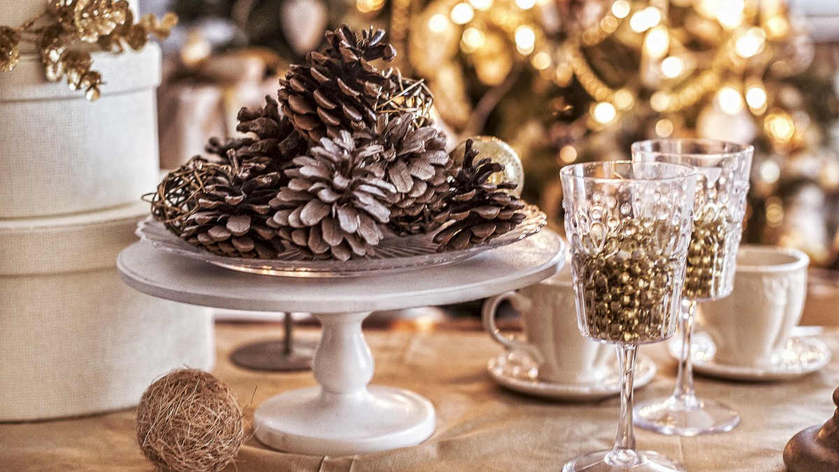 Festive table decor with pine cones on a stand, wine glasses, a string ball, and a blurred Christmas tree background with lights.