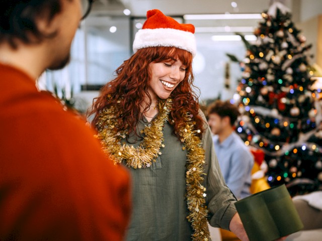 A woman with red hair wears a Santa hat and tinsel while smiling, with a decorated Christmas tree in the background at a festive gathering.