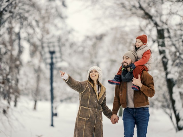 A happy family enjoys a snowy walk outdoors, with the dad carrying their child on his shoulders as the mom points at something in the distance.