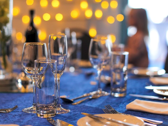 A formal dining table set with various glasses, plates, and cutlery, illuminated by warm, out-of-focus lights in the background.