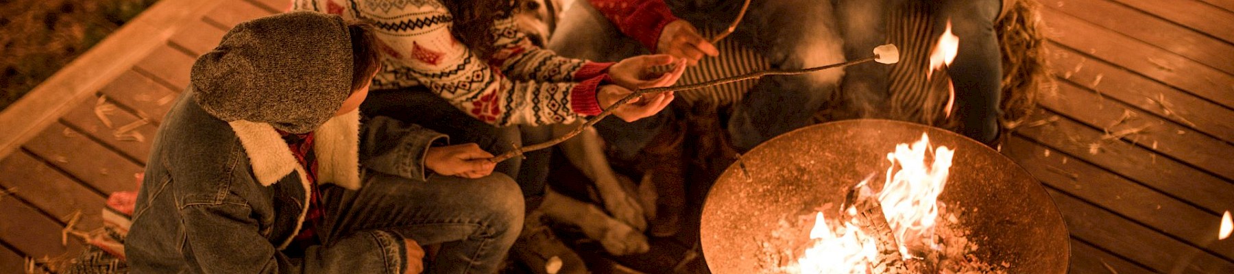 Four people in cozy sweaters roast marshmallows over a firepit on a wooden deck, surrounded by string lights, enjoying a warm evening.