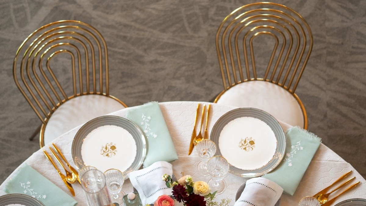 A round table set for a elegant meal with gold-rimmed plates, matching napkins, floral centerpiece, and stylish chairs, ready for guests.