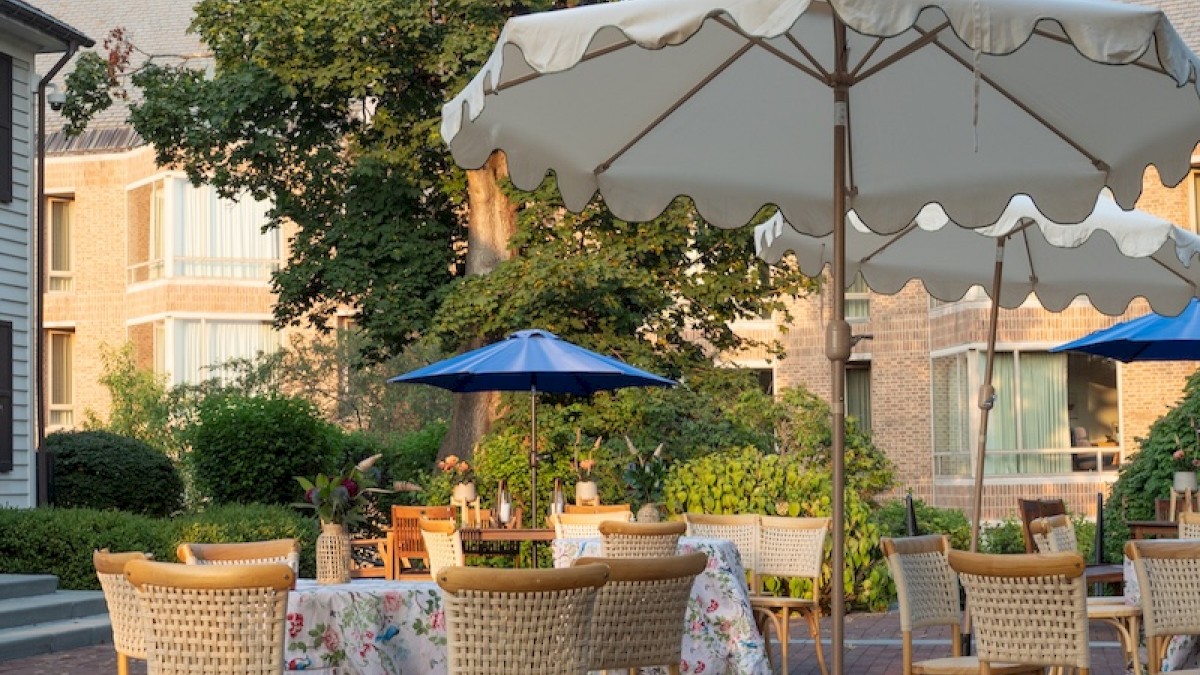 A quiet outdoor café with wicker chairs and checkered tables under large umbrellas, set on a brick patio near a green tree and buildings.
