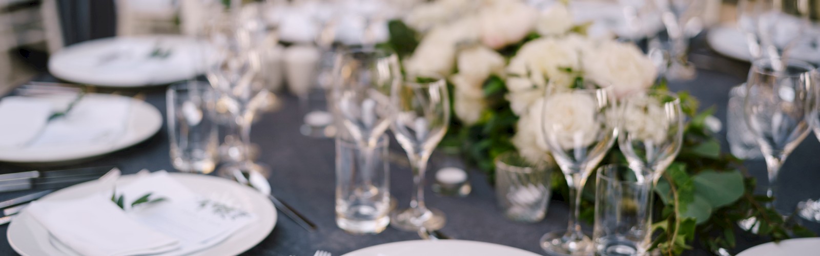 A table is elegantly set with white plates, glassware, and floral centerpieces, surrounded by white chairs, creating a formal dining ambiance.