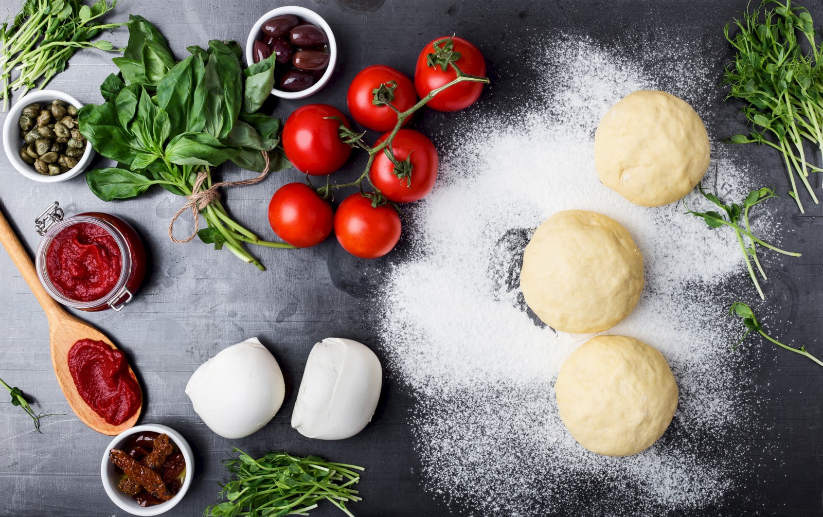 Pizza-making ingredients: dough balls, tomatoes, mozzarella, basil, spices, olives, capers, and tomato paste on a dark surface.