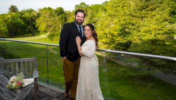 A couple is posing on a balcony with a scenic green landscape in the background, accompanied by a wooden bench and a bouquet.