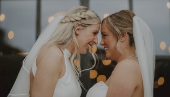 Two brides in white dresses and veils stand close, touching foreheads and smiling warmly against a softly lit background.