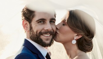 A couple poses lovingly under a veil; the woman kisses the man's cheek while he smiles warmly at the camera.