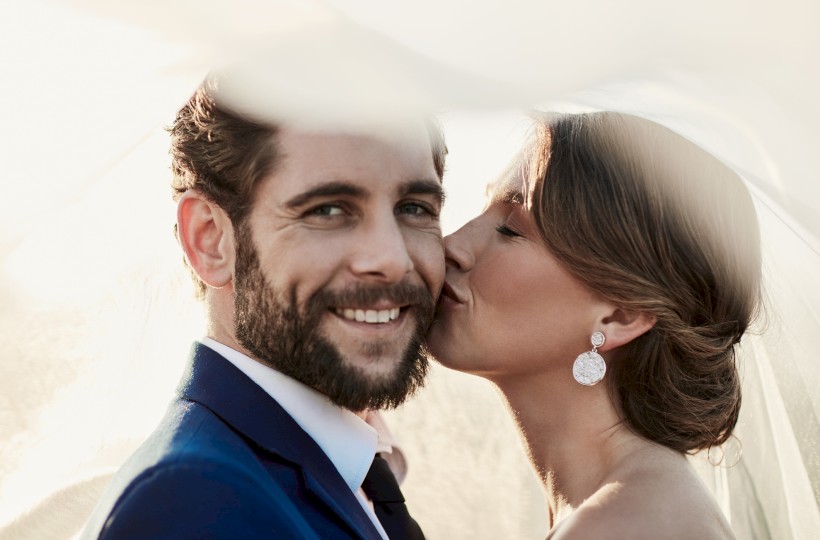 A couple poses lovingly under a veil; the woman kisses the man's cheek while he smiles warmly at the camera.