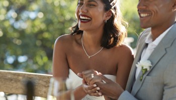 A happy couple is sitting together outdoors, holding hands and smiling, with one wearing a white dress and the other a light gray suit.
