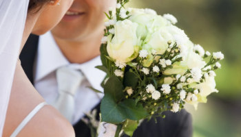 A bride and groom are close together; she's holding a bouquet of white roses and small white flowers.