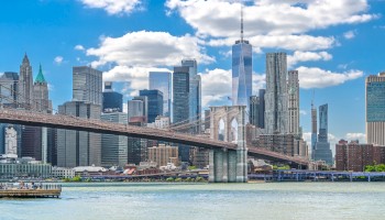 The image shows the Brooklyn Bridge with the Manhattan skyline in the background, featuring tall skyscrapers and a partly cloudy sky.