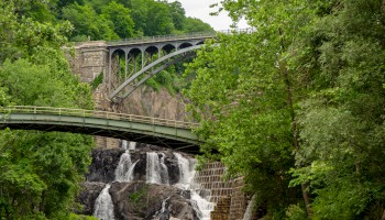The image shows two bridges over a waterfall surrounded by lush greenery, with a rocky foreground and trees framing the scene.