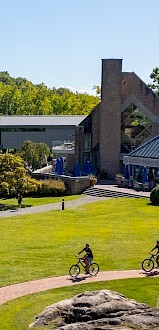 Two people cycle on a path through a green lawn, with a modern building and trees in the background.