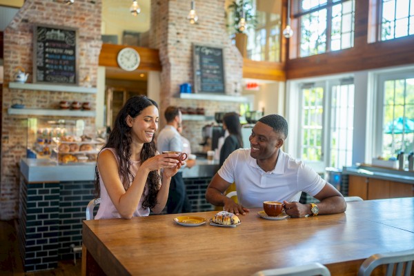 Two people enjoy coffee and pastries at a wooden table in a cozy café with brick walls and a menu chalkboard.