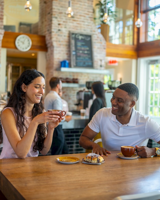 Two people enjoy coffee and pastries at a wooden table in a cozy café with brick walls and a menu chalkboard.