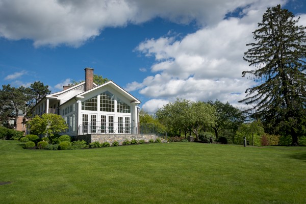A beautiful house with large windows sits amidst a lush green lawn, surrounded by trees under a partly cloudy sky.