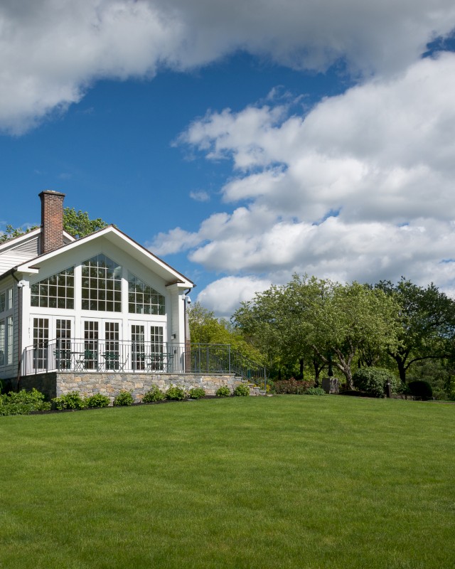 A beautiful house with large windows sits amidst a lush green lawn, surrounded by trees under a partly cloudy sky.