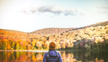 A person in a jacket and hat stands by a lake, admiring a scenic view of autumn trees and hills under a partly cloudy sky.