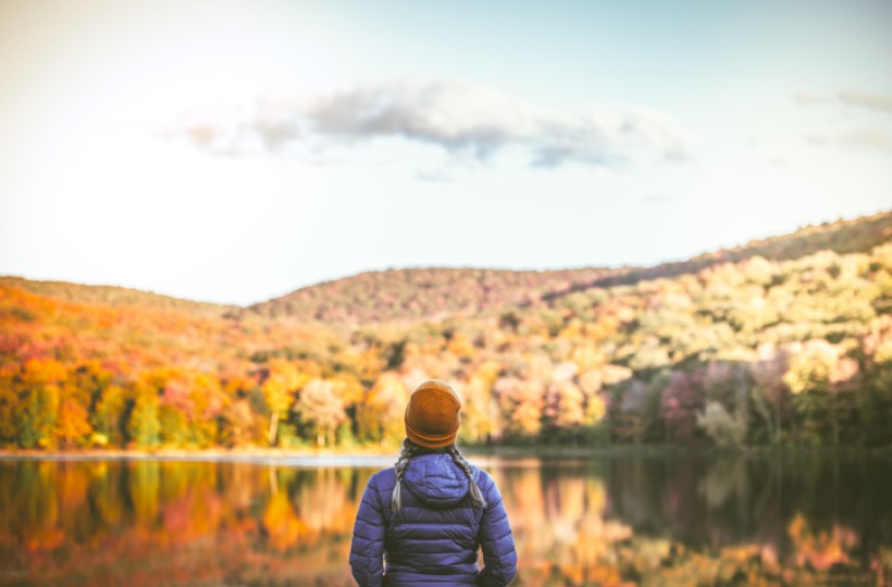 A person in a jacket and hat stands by a lake, admiring a scenic view of autumn trees and hills under a partly cloudy sky.