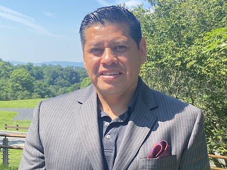 A person in a suit is standing outdoors in a scenic area with trees and hills in the background, under a clear blue sky.