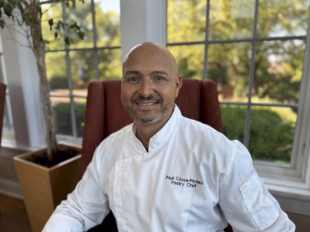 A person in a white chef's coat sits indoors, smiling. Behind them is a large window with greenery outside, and a potted plant is nearby.