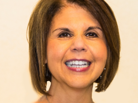 A woman with short brown hair is smiling, wearing a patterned blouse and a necklace, and standing against a plain background.
