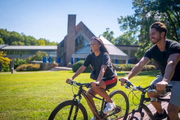 Two people ride bikes on a sunny lawn near a brick building, enjoying a daytime bike ride. (End with a period.)