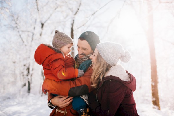 A family outdoors in a snowy forest: a smiling mother and father hold their toddler, all bundled in winter coats and hats, warm together.