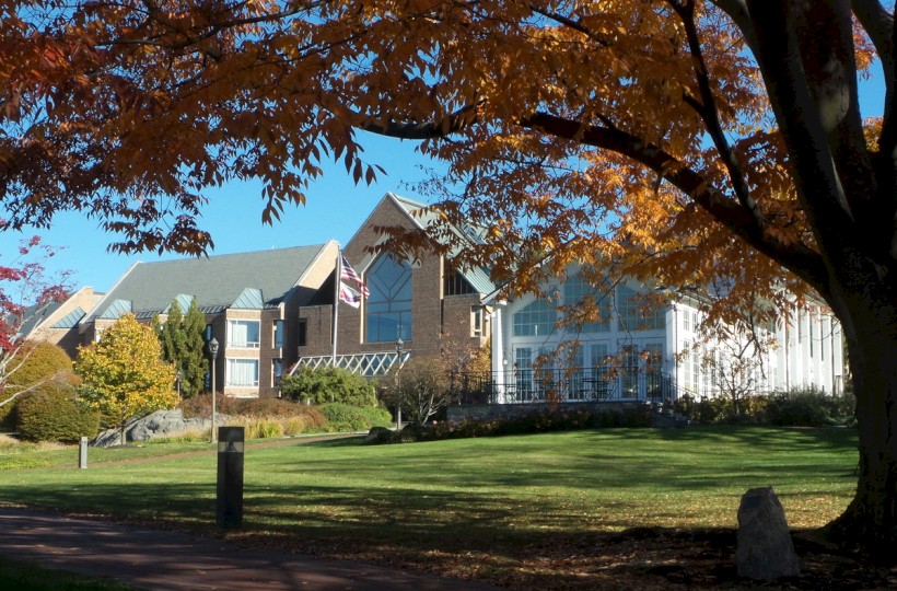 A scenic view of a modern building with large windows, surrounded by autumn trees, a well-maintained lawn, and a bright blue sky.