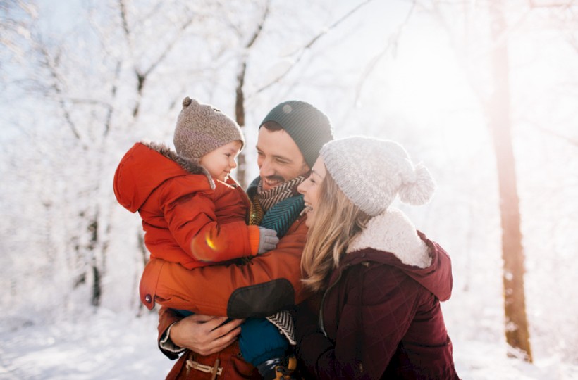 A family outdoors in a snowy forest: a mother and father smiling at their child in warm jackets and knitted hats, bright sunlight glow.