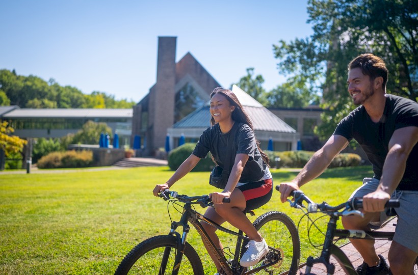 Two people ride bicycles on a sunny, grassy campus with a brick building in the background, enjoying a daytime outdoor ride.