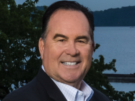 A person in a suit is smiling outdoors with trees and a body of water in the background.