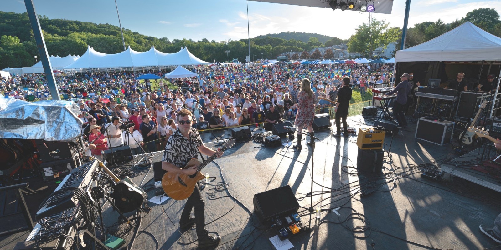 A band performs on a stage at an outdoor event with a large audience gathered under white tents on a sunny day.