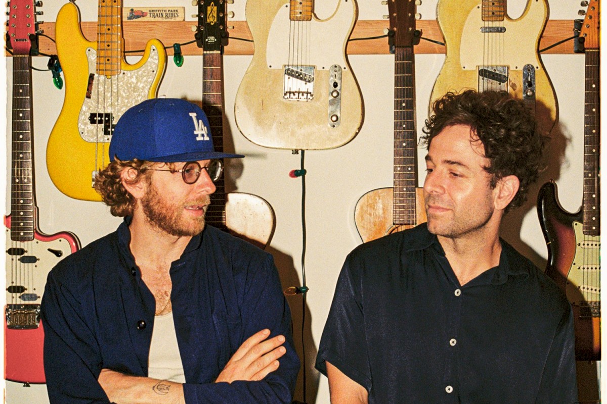 Two people are in front of a wall with electric guitars. One wears a blue cap and glasses, the other has a black shirt.