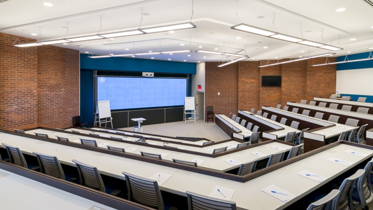 A modern lecture hall with tiered seating, desks, chairs, a large screen at the front, and brick walls.