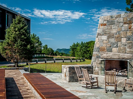 Outdoor patio with wooden chairs and a large stone fireplace, surrounded by trees and buildings under a blue sky with scattered clouds.