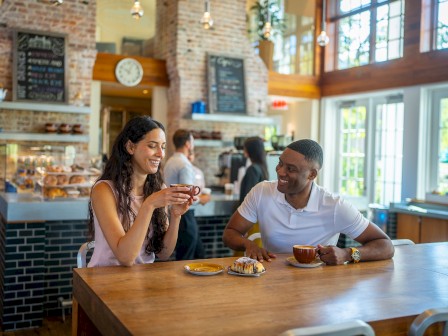 A man and woman enjoy coffee and pastries at a cafe with a brick interior, large windows, and other people in the background.