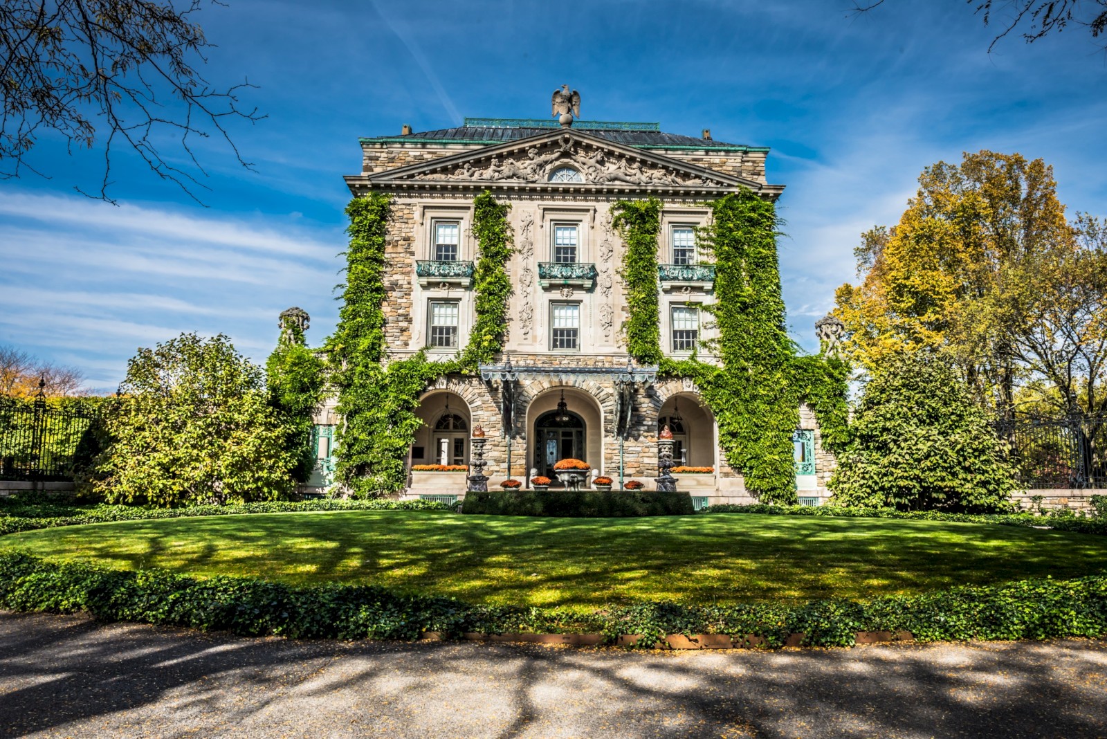 A grand stone house with ivy-covered walls, surrounded by lush green lawns and trees under a blue sky.